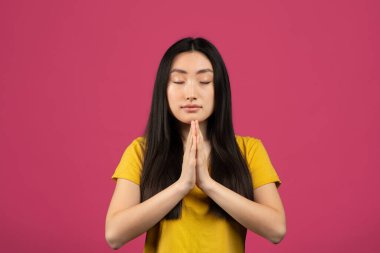 Dua et ve meditasyon yap. Çinli bir kadının portresi avuçlarını bir arada tutup dua ediyor, meditasyon yapıyor, sakin ve huzurlu bir yüz ifadesi takınıyor, pembe arka plan, stüdyo çekimi.