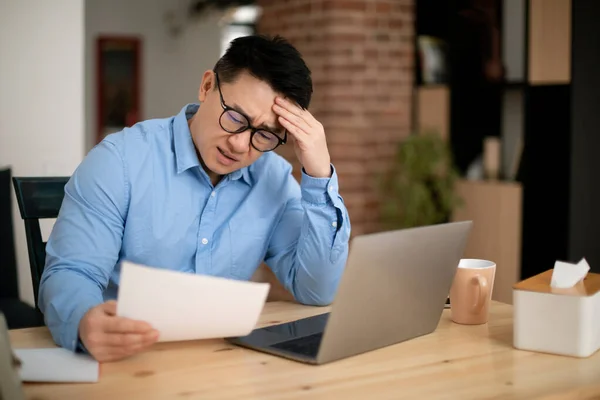 Confused asian mature man sitting in front of laptop, reading papers and touching his head, copy space. Korean businessman working with documents while working from home