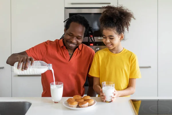 アフリカ系アメリカ人の父と彼の十代の娘は キッチンでお菓子を持っています ハッピーブラックお父さんとかわいい女性の子供は自家製のペストリーを食べ 楽しみを持っています お父さんはボトルからガラスにミルクを注ぎます — ストック写真