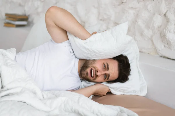 Tired angry sad young caucasian guy lies on bed, covers his ears with pillow, suffers from noisy neighbors, stress and depression in bedroom interior. Disease, human emotions and insomnia at home