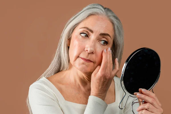 Serious elderly caucasian lady with gray hair looks in mirror, touches wrinkles, applies cream, basic on face isolated on brown background, studio. Skin care, nude makeup, daily procedures for beauty