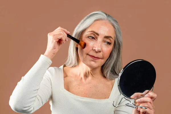 Smiling elderly caucasian woman with gray hair applying blush on her cheeks, looks in mirror isolated on brown background, studio. Beauty care, makeup and cosmetics, daily procedures, ad and offer
