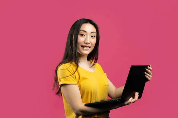 Remote work. Happy chinese female freelancer using laptop, enjoying distance job, standing over pink studio background, copy space. Lady browsing internet on pc computer