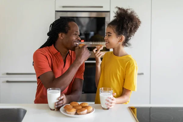幸せな黒人の父と十代の娘は家でお菓子を食べ キッチンで楽しみを持っています 陽気なアフリカ系アメリカ人の家族のお父さんと女性は自家製マフィンを楽しんで ミルクを飲みます — ストック写真
