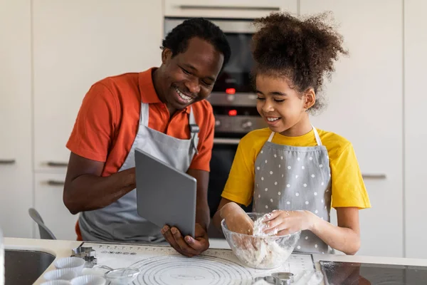 Smiling Black Dad And Preteen Daughter Baking And Using Digital Tablet In Kitchen, Happy African American Father And Female Child Checking Ingredients Online, Searching For Cookies Recipe