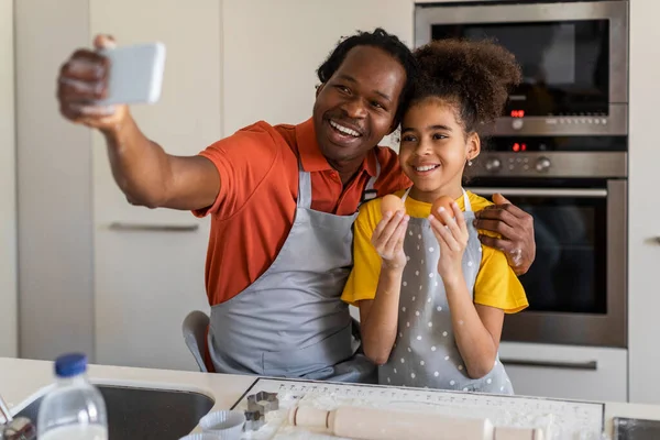 Family Fun. Joyful Black Father And Daughter Taking Selfie On Smartphone In Kitchen, Happy African American Dad And Preteen Female Child Posing At Mobile Phone Camera While Baking At Home