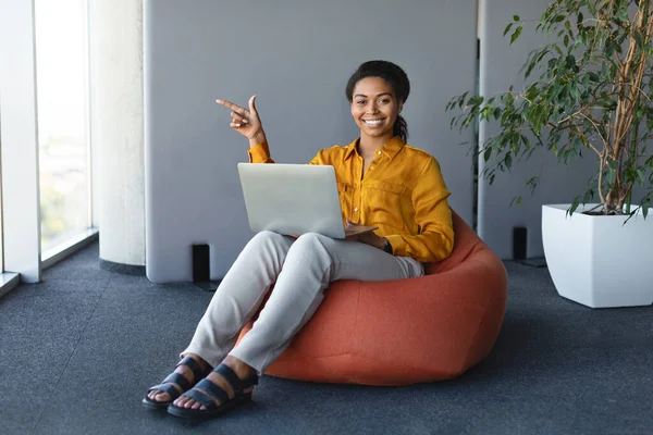 Happy Black Businesswoman Sitting Beanbag Chair Laptop Pointing Finger Aside — Foto Stock