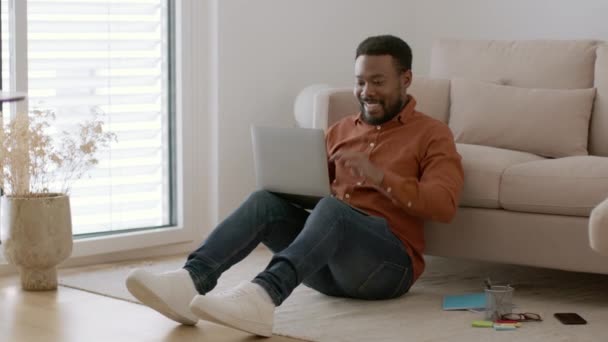 オンライン達成 若い屈託のないアフリカ系アメリカ人の男性フリーランスがラップトップで入力し 受賞歴のある電子メールと笑いを読み 自宅で床に座って 撮影を追跡し スローモーション フリースペース — ストック動画