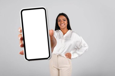 Beyaz bluzlu, güler yüzlü siyah kadın boş ekranlı telefon gösterisi gri arka planda ve stüdyoda izole edilmiş yeni bir uygulama öneriyor. İş, iş, öğrenim, online reklam ve teklif için araç