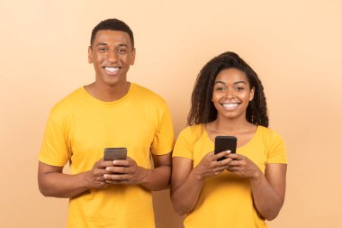 Mutlu siyah çift portresi cep telefonu kullanıyor ve kameraya gülümsüyor, sarı stüdyo arka planında duruyor. Yeni uygulama ve internet kullanan Afrikalı Amerikalı eşler