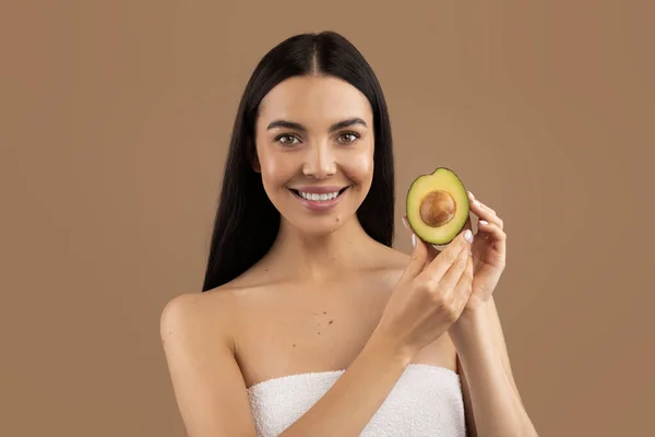Natural skin care. Portrait of beautiful brunette half-naked adult woman holding avocado half in her hand, lady looking at camera and gently smiling. Isolated on beige studio background, copy space