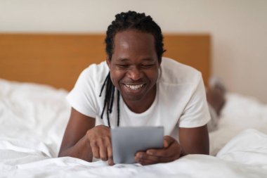 Mutlu siyah adam yatakta dinlenirken modern dijital tablet kullanıyor, yakışıklı Afrikalı Amerikalı adam yatak odasında sekme bilgisayarla dinleniyor, online alışveriş yapıyor ya da interneti evde tarıyor.