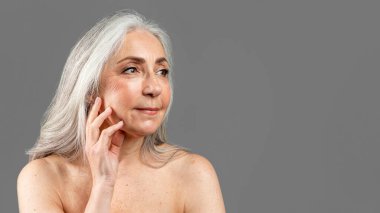 Kafkasyalı mutlu kadın elleriyle yüzüne dokunuyor, güzellik prosedürlerinin sonuçlarından zevk alıyor, boş alana bakıyor, gri arka planda, stüdyoda izole edilmiş. Tedavi, yaşlanma karşıtı cilt bakımı, reklam ve sunum
