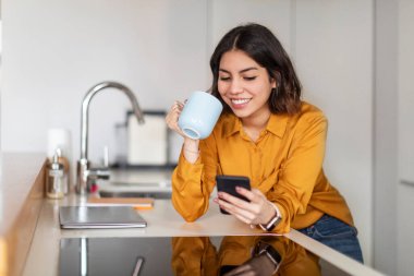 Smartphone 'da sosyal ağları tarayan ve evde kahve içen mutlu bir Arap kadını, çevrimiçi iletişim için cep telefonu kullanan gülümseyen genç bir Orta Doğulu kadın,