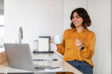 Gülümseyen Arap kadın bilgisayarla bilgisayar aracılığıyla arama yapıyor ve mutfakta kahve içiyor, genç ve mutlu bir Orta Doğulu kadın bilgisayar kamerasında konuşuyor, çevrimiçi iletişimin tadını çıkarıyor,
