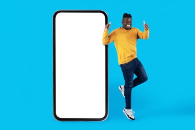 Harika bir teklif. Beyaz Ekranlı, Büyük Boş Akıllı Telefonun yanından atlayan ve Başparmağıyla hareket eden heyecanlı Afrikalı Amerikalı Adam Mobil Uygulamayı Öneriyor ya da Mavi Arkaplan Üzerinde Web Sitesi