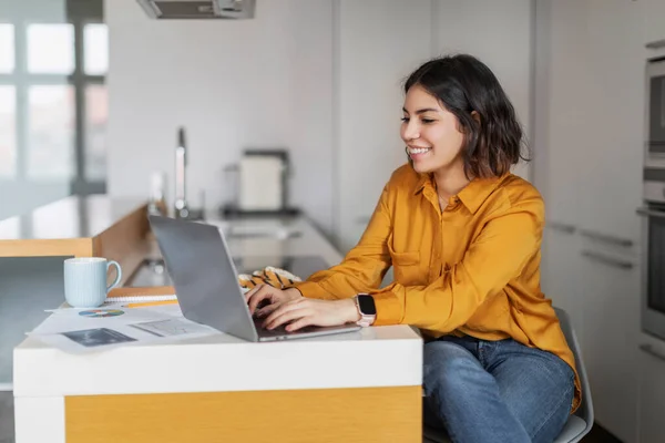 Remote Work. Smiling Young Arab Female Freelancer Working With Laptop In Kitchen Interior, Happy Millennial Middle Eastern Woman Using Computer At Home, Enjoying Online Job, Free Space