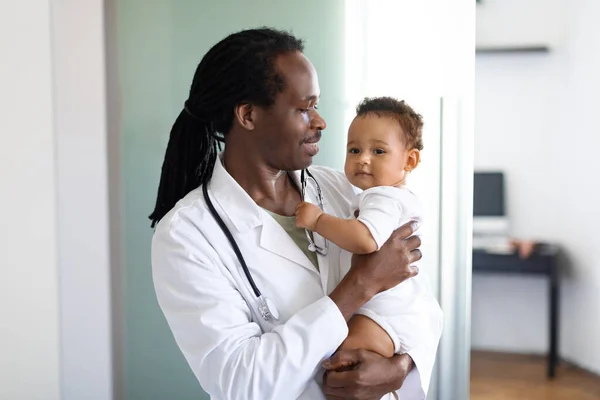Portrait Of Smiling Black Pediatrician Doctor Holding Cute Infant Baby In Arms, African American Pediatrist Man In Medical Uniform Bonding With Little Child During Appointment In Clinic, Copy Space