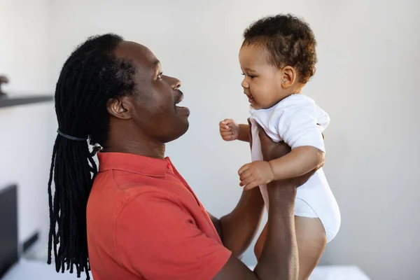 Portrait Of Young Black Father Chatting With Cute Little Baby At Home, Loving African American Dad Holding Adorable Infant Boy Or Girl In Arms, Enjoying Bonding With His Kid, Closeup Shot