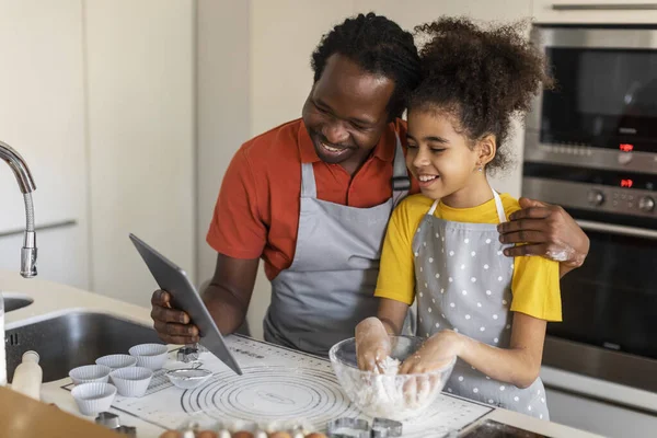 Black Father Using Digital Tablet While Baking With Daughter At Home, Happy African American Dad And Preteen Female Child Checking Muffins Recipe Online, Enjoying Cooking Pastry Together, Free Space