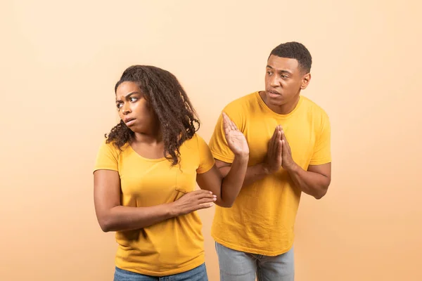 Black husband trying to talk and reconcile with offended wife, standing over yellow studio background. Marital relationship problems, family conflicts and divorce concept