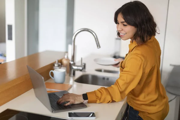 Young Arab Female Using Laptop Computer Washing Dishes Kitchen Smiling — Zdjęcie stockowe