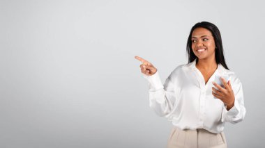 Happy pretty millennial black female in white blouse typing on smartphone, showing finger on free space isolated on gray background. Ad and offer, gadget for business, app for work, study and startup