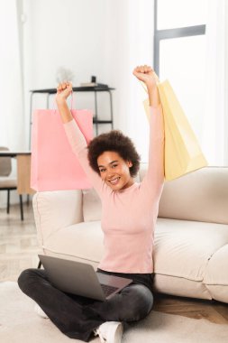 Glad young mixed race lady shopaholic sits on floor with laptop, raises hands up with packages from store, celebrating success in living room interior. Online shopping at home, huge sale and cashback