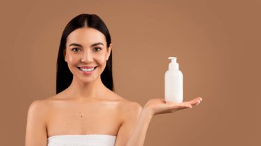 Attractive millennial long-haired brunette half-naked woman with perfect body and young smooth skin recommending body cream or lotion, holding white bottle on palm and smiling, beige studio background
