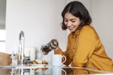 Closeup Shot Of Young Smiling Arab Woman Pouring Morning Coffee To Cup, Beautiful Happy Middle Eastern Female Holding French Press And Preparing Caffeine Drink White Resting In Modern Kitchen At Home