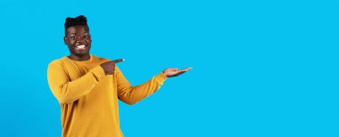 Try This. Happy Young Black Male Pointing At His Empty Palm, Smiling Millennial African American Man Demonstrating Free Place For Advertisement Or Offer Design Over Blue Background, Panorama