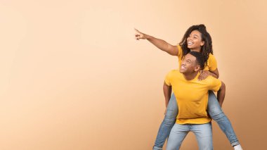 Excited african american couple having fun, lady piggybacking guy and pointing finger at copy space on yellow background, demonstrating place for advert, panorama