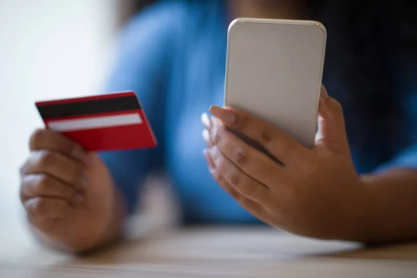 Cropped shot of dark-skinned chubby woman in blue shirt sitting at desk indoors, holding modern cell phone and red bank card. Female customer banking or shopping online