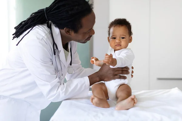 Smiling Pediatrician Doctor Talking To Black Infant Baby During Appointment In Clinic, Professional Medical Worker In Uniform Making Health Check-Up To Adorable Little Boy Or Girl In Hospital