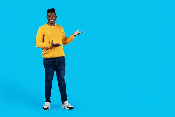 Look There. Happy Young Black Man Pointing Aside At Copy Space With Two Palms, Cheerful African American Guy Demonstrating Free Place For Advertisement While Standing Isolated On Blue Background