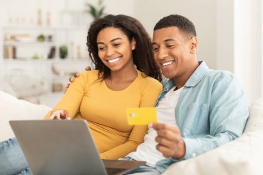 Cheerful young african american guy and woman use credit card and laptop, check bank financial account, sit on sofa in living room interior. Sale for shopaholics, online shopping, ad and offer at home