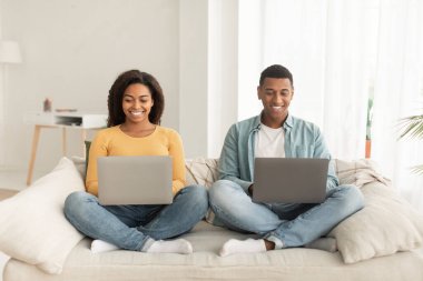 Cheerful young african american guy and lady with laptops work, surfing in internet, sit on sofa in minimalist living room interior. New normal, business, freelance and free time with device at home