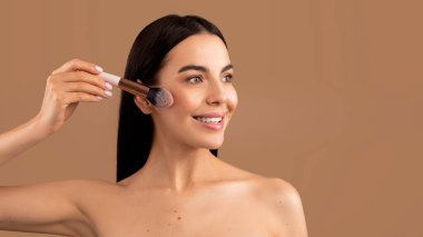 Happy smiling attractive brunette half-naked young woman with long hair holding brush, applying blush and looking at copy space over beige studio background, makeup concept, panorama