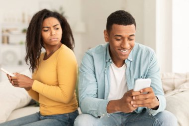 Unhappy jealous young african american female with smartphone looks at male phone in room interior. Addiction from device, online games and social networks, relationship problems, cheating at home