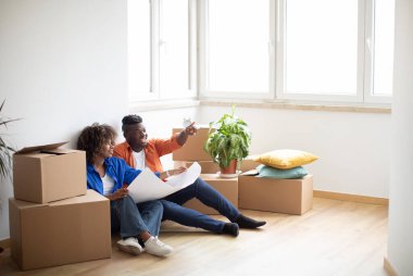 Black Young Couple Holding Blueprints, Planning Design Of Their New Home Together After Moving, Happy African American Spouses Sitting On Floor Among Cardboard Boxes And Discussing House Furniture
