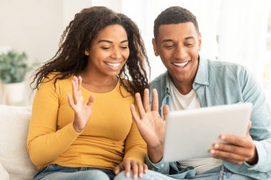 Cheerful young african american guy and lady waving hands at tablet webcam, greeting, say hello on sofa in living room interior, close up. Application for communication and meeting, new normal at home
