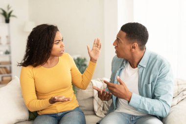 Angry sad emotional young african american lady and male screaming, gesturing, sitting on sofa in living room interior. Quarrel, family relationship problems, divorce and crisis at home due covid-19