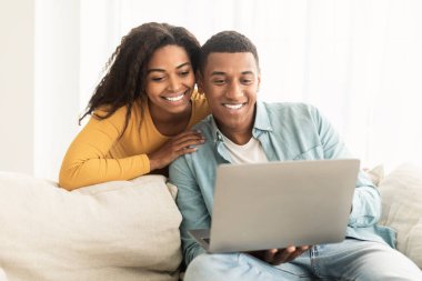 New normal and meeting remote. Glad young african american family look at computer webcam, greeting on sofa in living room interior. Video call, app for communication, funny video on device at home