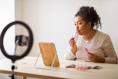 Cool young hispanic woman plus size influencer sitting at vanity table in front of small mirror, streaming from home while applying lipstick, using phone, famous beauty blogger recommending lip gloss
