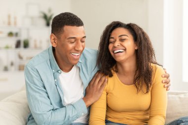 Laughing handsome young african american woman and guy hug, have fun in free time, sitting on sofa in living room interior, close up. Love, family relationship, support and care at own home, covid-19