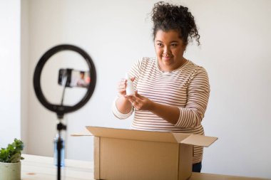 Cute pretty mixed race young overweight woman beauty blogger showing followers face care product white bottle, lady influencer streaming via cell phone while unpacking cosmetics skin care at home