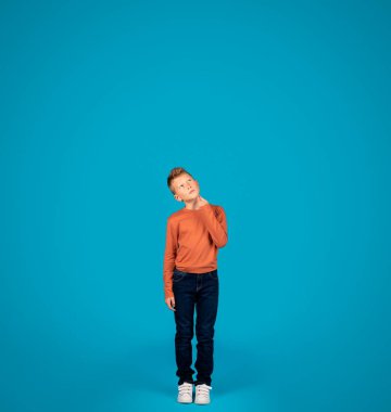 Thoughtful Preteen Boy Looking Up While Standing Over Blue Studio Background, Pensive Caucasian Male Child Touching Chin, Thinking And Considering Options, Full Length Shot, Copy Space