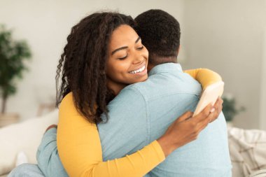 Cheerful millennial african american lady hugging man and chatting in smartphone, ordering goods in home interior. Addiction from device and social networks, problems in relationships, ad and offer