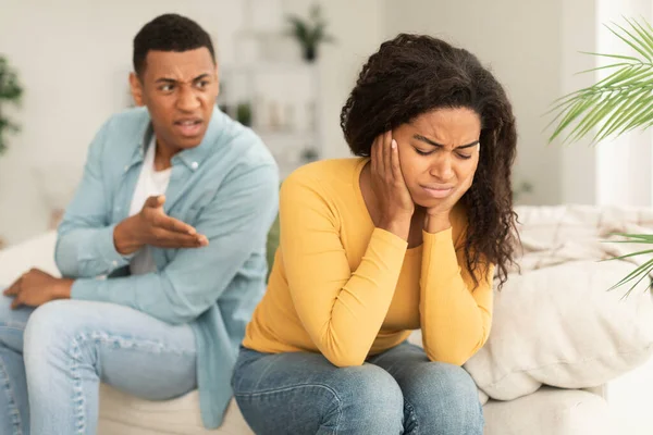 Sad crying young african american woman ignores angry aggressive screaming guy sit on sofa, think about breakup in room interior. Scandal quarrel, relationship problems, crisis and divorce at home