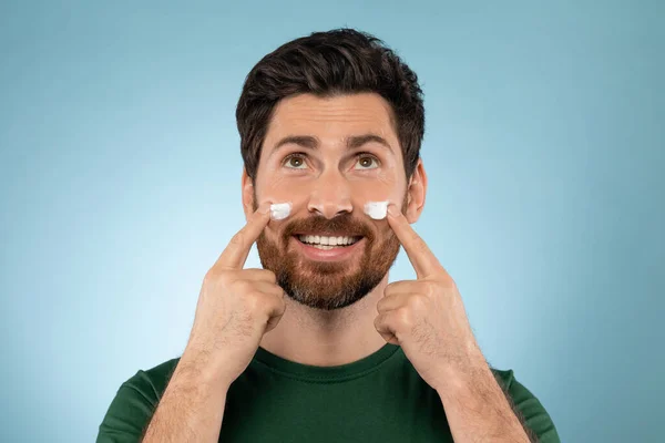 Bearded handsome man applying face cream on cheeks, standing on blue studio background, looking and smiling at camera. Good looking guy using beauty products for his face
